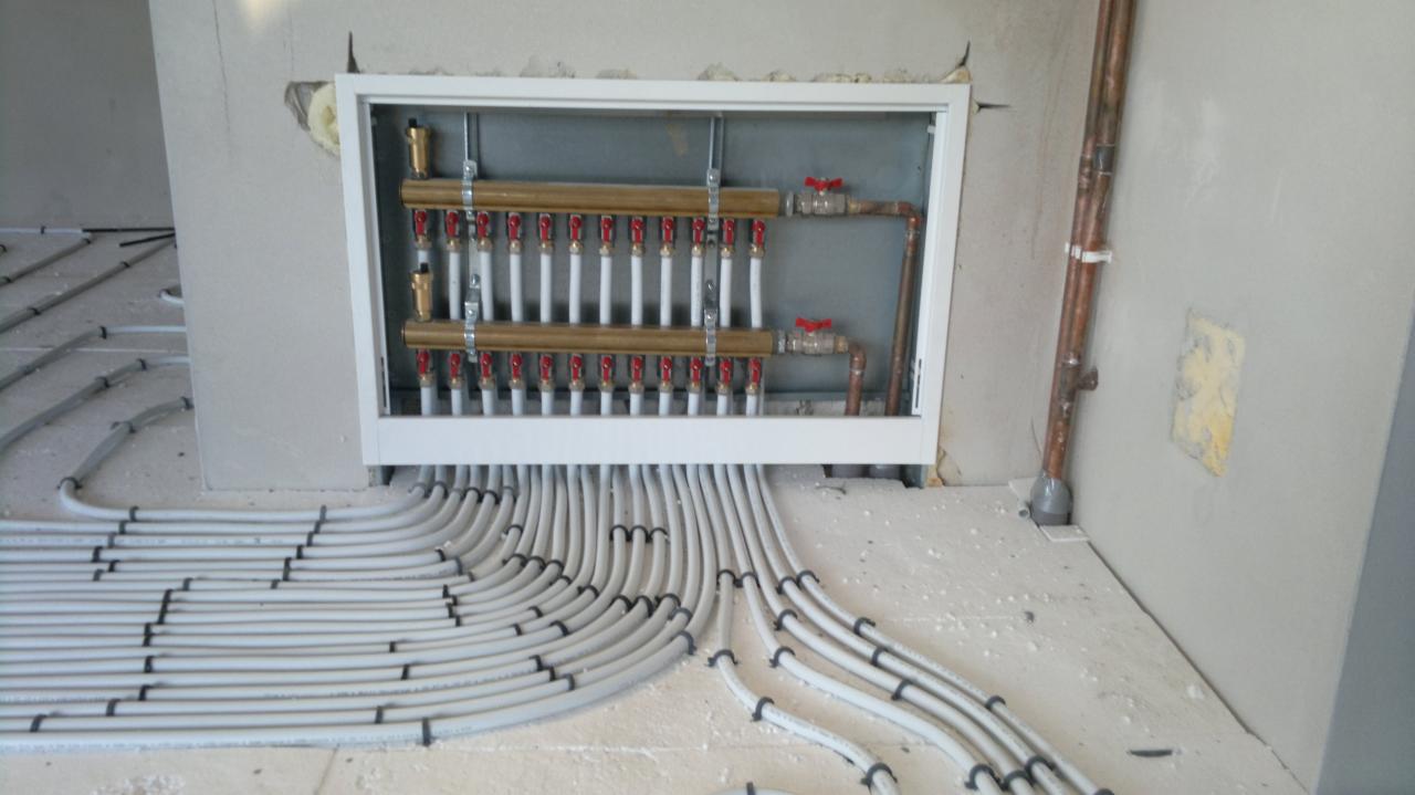 Rozdzielacz ogrzewania podłogowego z licznymi pętlami rur na jasnym podłożu, widoczne zawory z czerwonymi pokrętłami i miedziana rura przy ścianie.