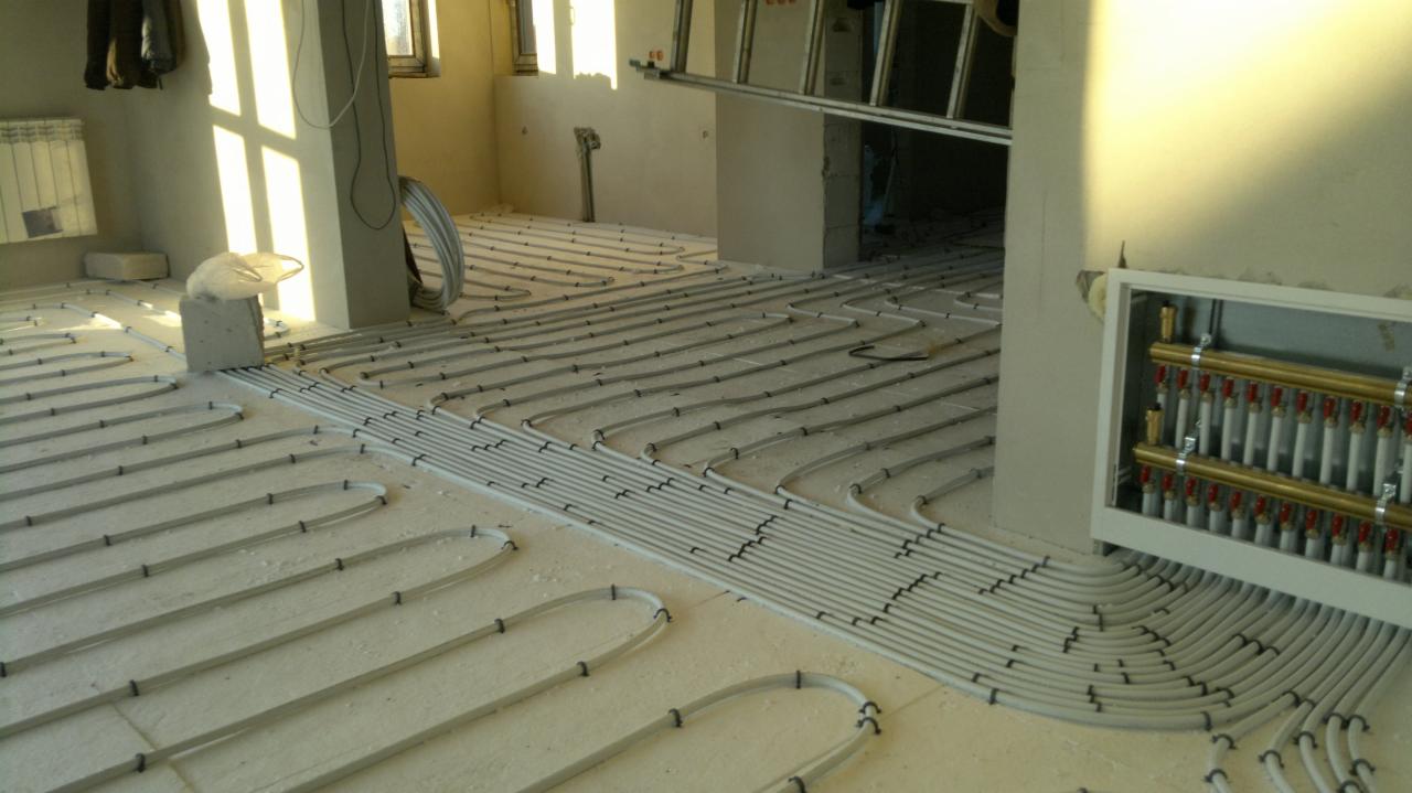 Rozłożone rury ogrzewania podłogowego na izolacji, z widocznym rozdzielaczem w białej obudowie i porannym światłem wpadającym przez okno.