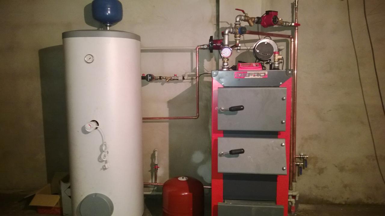 Zmontowany, dwudrzwiowy piec centralnego ogrzewania marki DEFRO, obok biały bojler z niebieskim zbiornikiem wyrównawczym, instalacja z miedzianych rur.