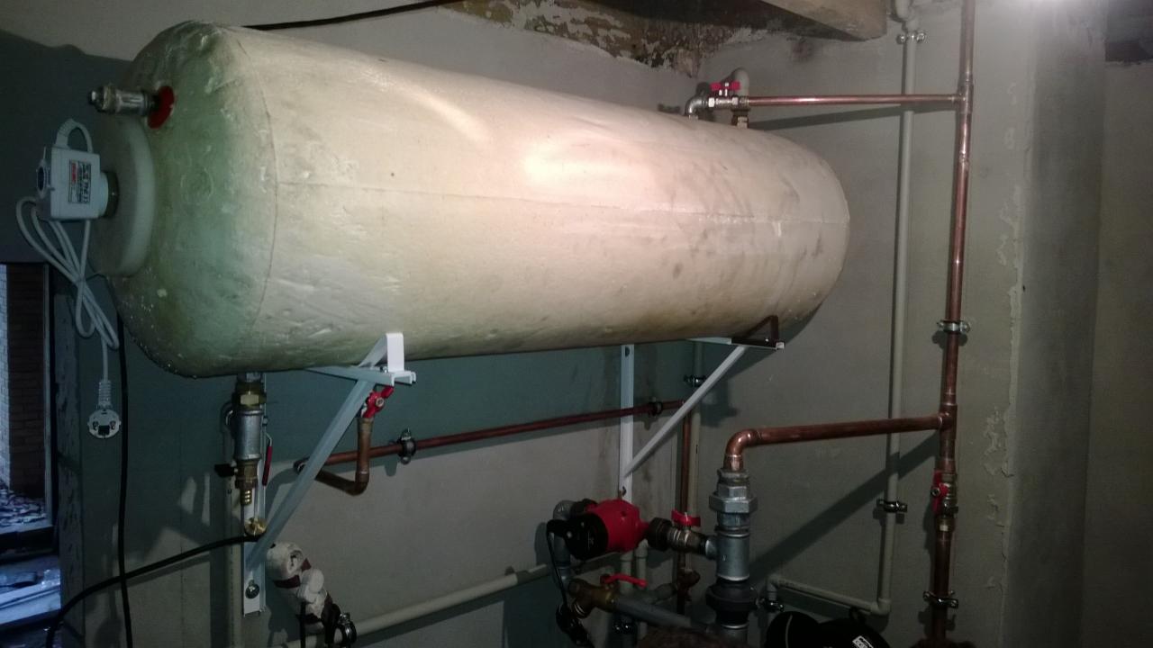 Poziomy zbiornik akumulacyjny ciepła, zamontowany na ścianie na stalowych wspornikach, z podłączonymi miedzianymi rurami i pompą obiegową w kotłowni.