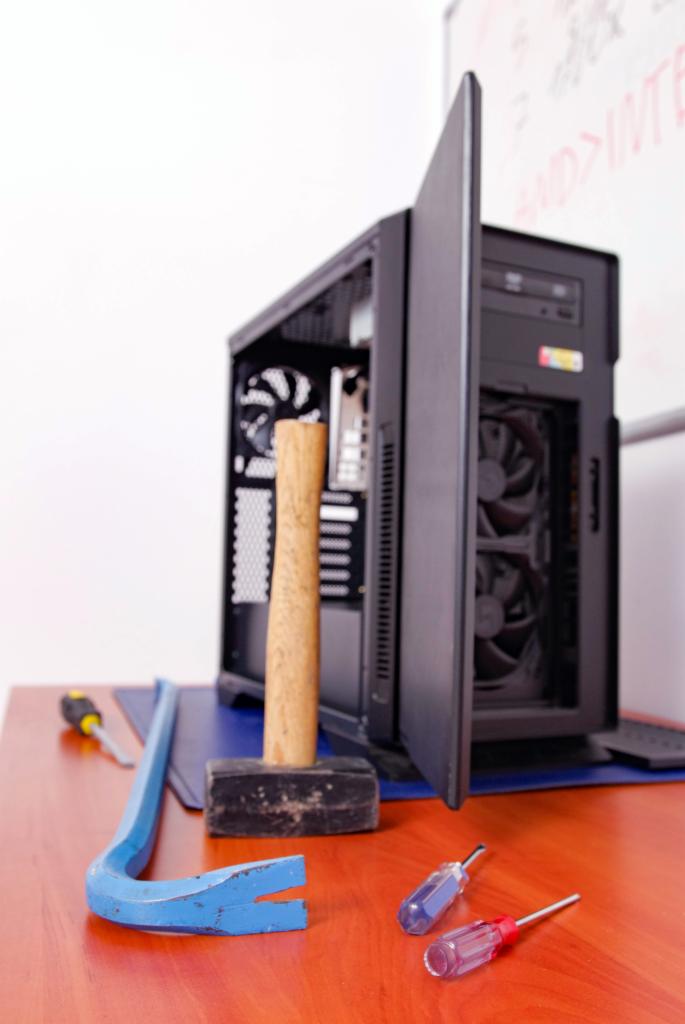 Otwarta obudowa komputera stacjonarnego z widocznym wentylatorem i płytą główną, obok leżą narzędzia: młotek, łom i dwa śrubokręty z przezroczystymi uchwytami na drewnianym biurku z niebieską...