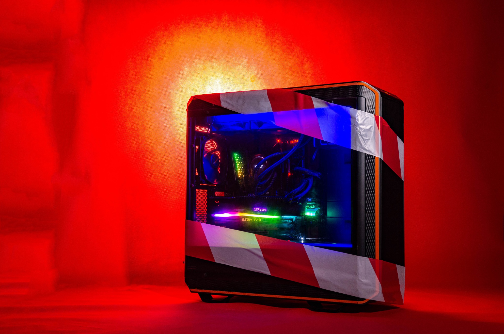 Komputer stacjonarny z przezroczystą obudową, owinięty taśmą ostrzegawczą w biało-czerwone paski, podświetlony na niebiesko i zielono, na czerwonym tle.