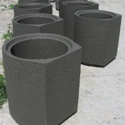 Szereg szarych, betonowych donic o geometrycznym kształcie, ustawionych na szarym podłożu z drobnymi kamieniami i kępkami zieleni.