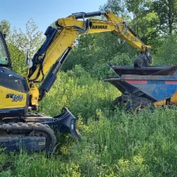 Żółto-czarna mini koparka Yanmar SV100 na gąsienicach, stojąca na zarośniętym terenie, ładuje urobek do niebiesko-żółtej wywrotki Barford SX6000, w tle widoczne drzewa i fragment drogi.