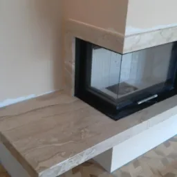 Narożny wkład kominkowy obudowany beżowym kamieniem z wyraźnym wzorem żyłkowania; widoczne fragmenty wykończenia ścian i parkietu.