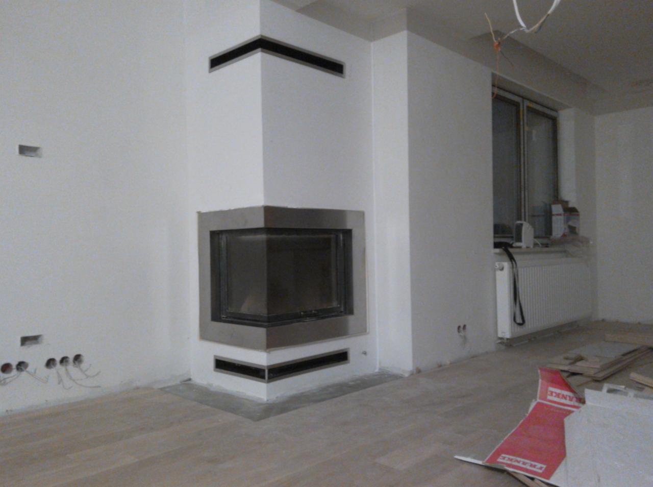 Narożny wkład kominkowy z metalowym obramowaniem, w trakcie montażu, widoczne niedokończone ściany, instalacja elektryczna i przygotowane podłoże pod podłogę.