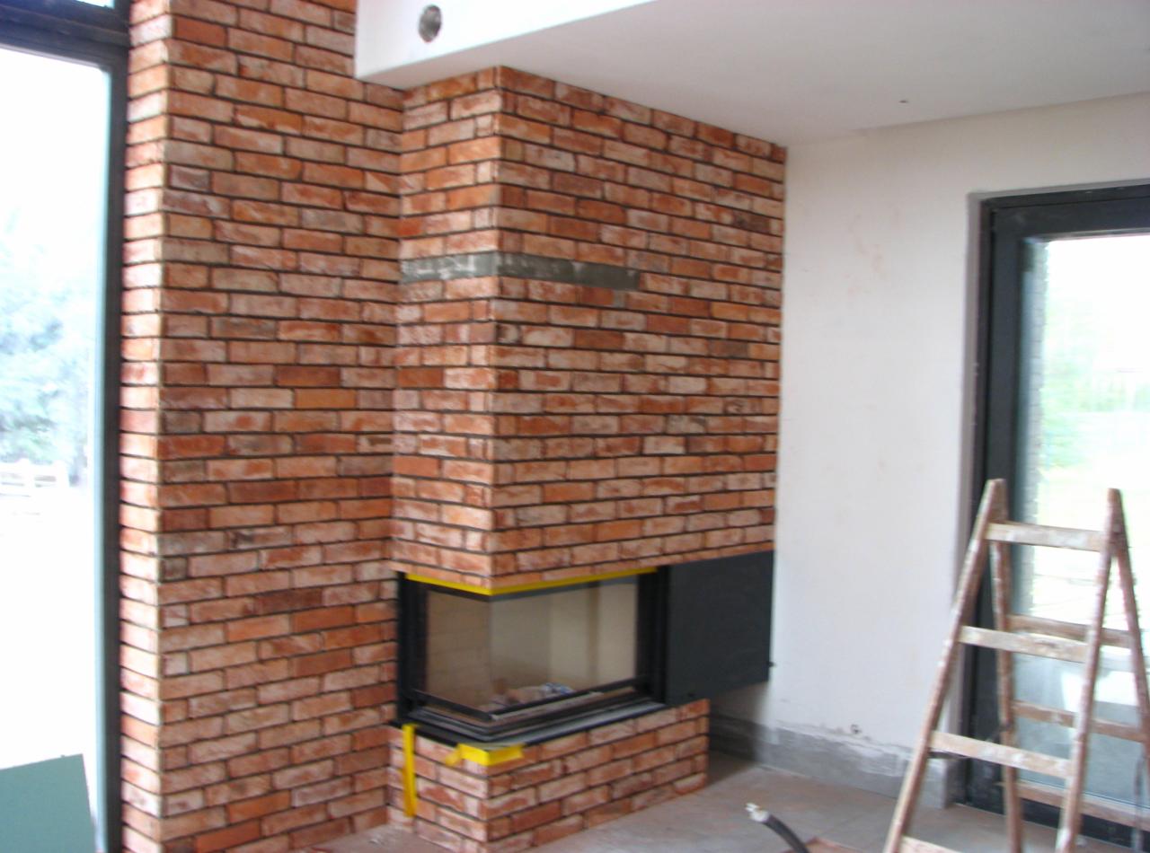 Murowany kominek z cegły klinkierowej w trakcie wykończenia, widoczne zabezpieczenia taśmą malarską, drabina oparta o ścianę obok.