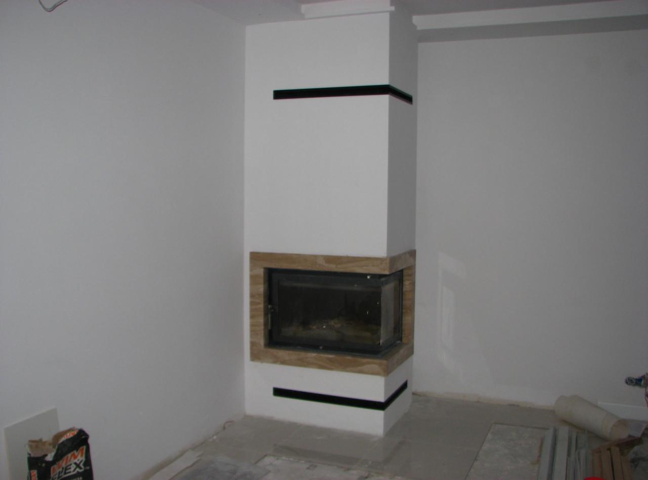 Nowoczesny kominek narożny z obudową z jasnego kamienia i czarnymi akcentami, w trakcie montażu, widoczny worek z zaprawą klejową.