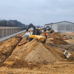 ANDRO BRUKARSTWO Robert Pawlak, Mariusz Pawlak s.c. - Żółta koparka Mecalac na placu budowy, widoczne wykopy pod rury, pracownicy w pomarańczowych kamizelkach, w tle budynek i drzewa. Prace ziemne w toku.