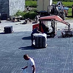 ANDRO BRUKARSTWO Robert Pawlak, Mariusz Pawlak s.c. - Prace brukarskie w toku: robotnicy układają szarą kostkę brukową na dużej powierzchni. W tle maszyny budowlane i palety kostki czekające na rozładunek.