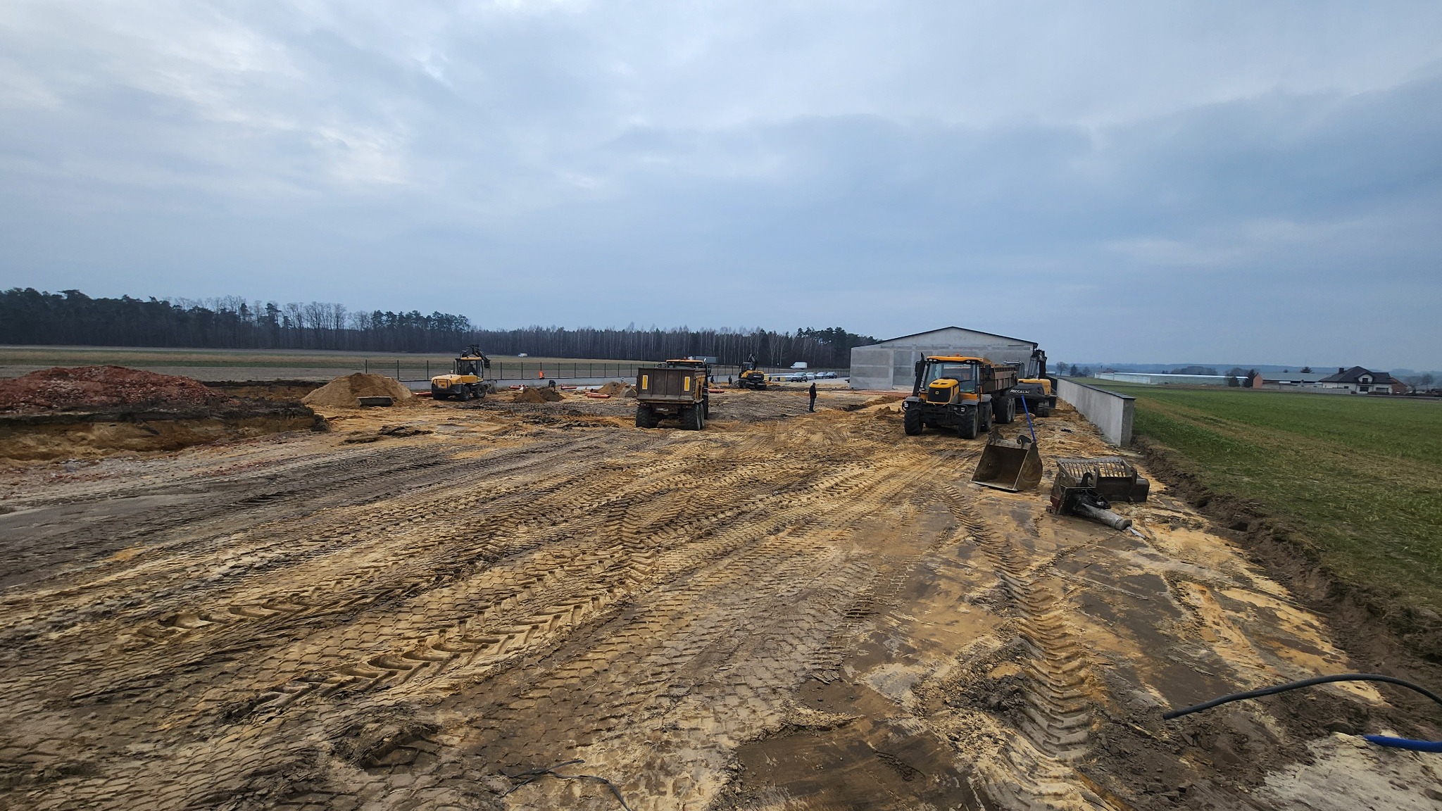 Szeroki kadr na teren budowy z ciężkim sprzętem: koparki, wywrotka. W tle budynek gospodarczy, łąka i las pod pochmurnym niebem. Ślady opon na ubitej ziemi.