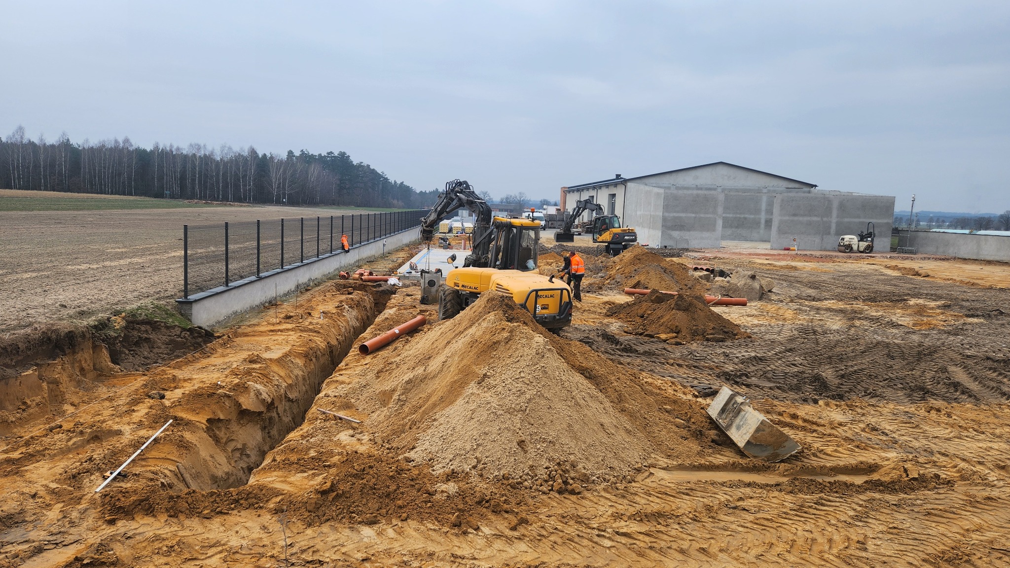 Żółta koparka Mecalac na placu budowy, widoczne wykopy pod rury, pracownicy w pomarańczowych kamizelkach, w tle budynek i drzewa. Prace ziemne w toku.