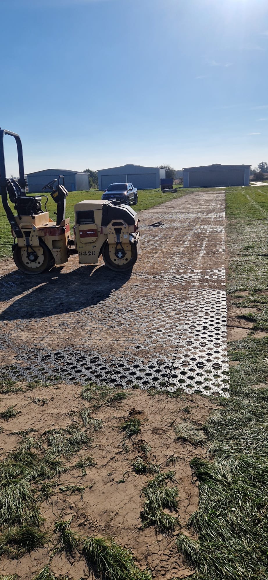 Żółty walec drogowy ubija plastikową kratkę trawnikową na gruncie. W tle budynki gospodarcze i niebo.