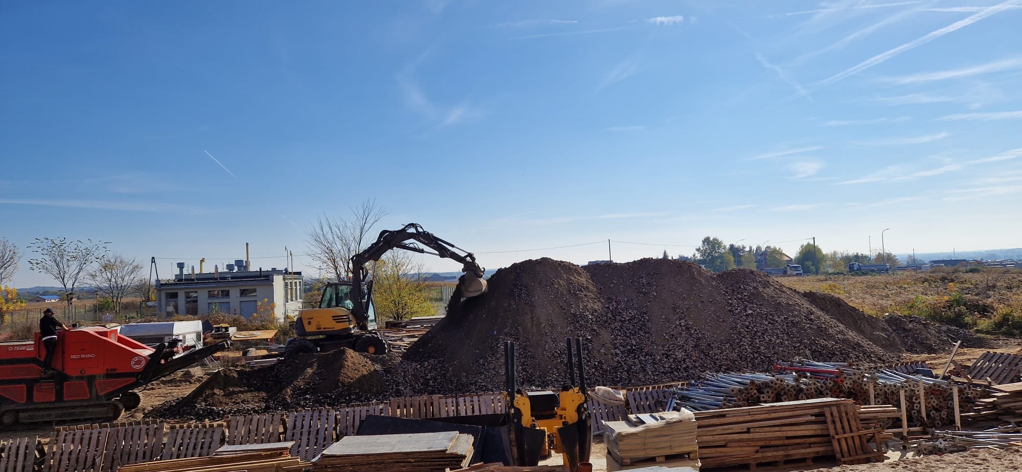 Żółta koparka nabiera ziemię na hałdę. W tle budynek, drzewa i niebo. Widoczne maszyny i materiały budowlane. Działka budowlana w trakcie prac ziemnych.