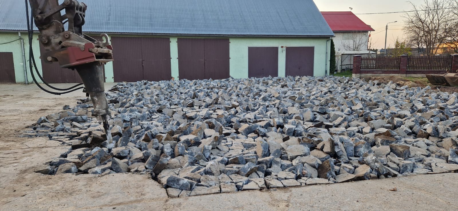 Kruszenie betonu na placu za pomocą młota pneumatycznego. W tle budynek gospodarczy z brązowymi bramami i fragment drugiego budynku z czerwonym dachem.