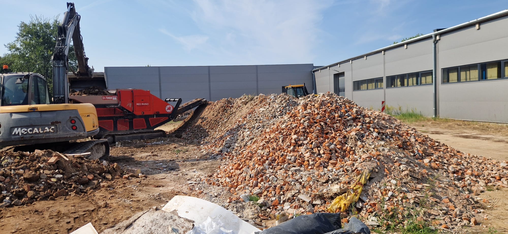 Żółta koparka Mecalac ładuje gruz do czerwonej kruszarki Red Rhino. W tle budynek przemysłowy i błękitne niebo.