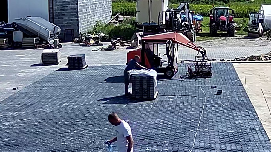 Prace brukarskie w toku: robotnicy układają szarą kostkę brukową na dużej powierzchni. W tle maszyny budowlane i palety kostki czekające na rozładunek.