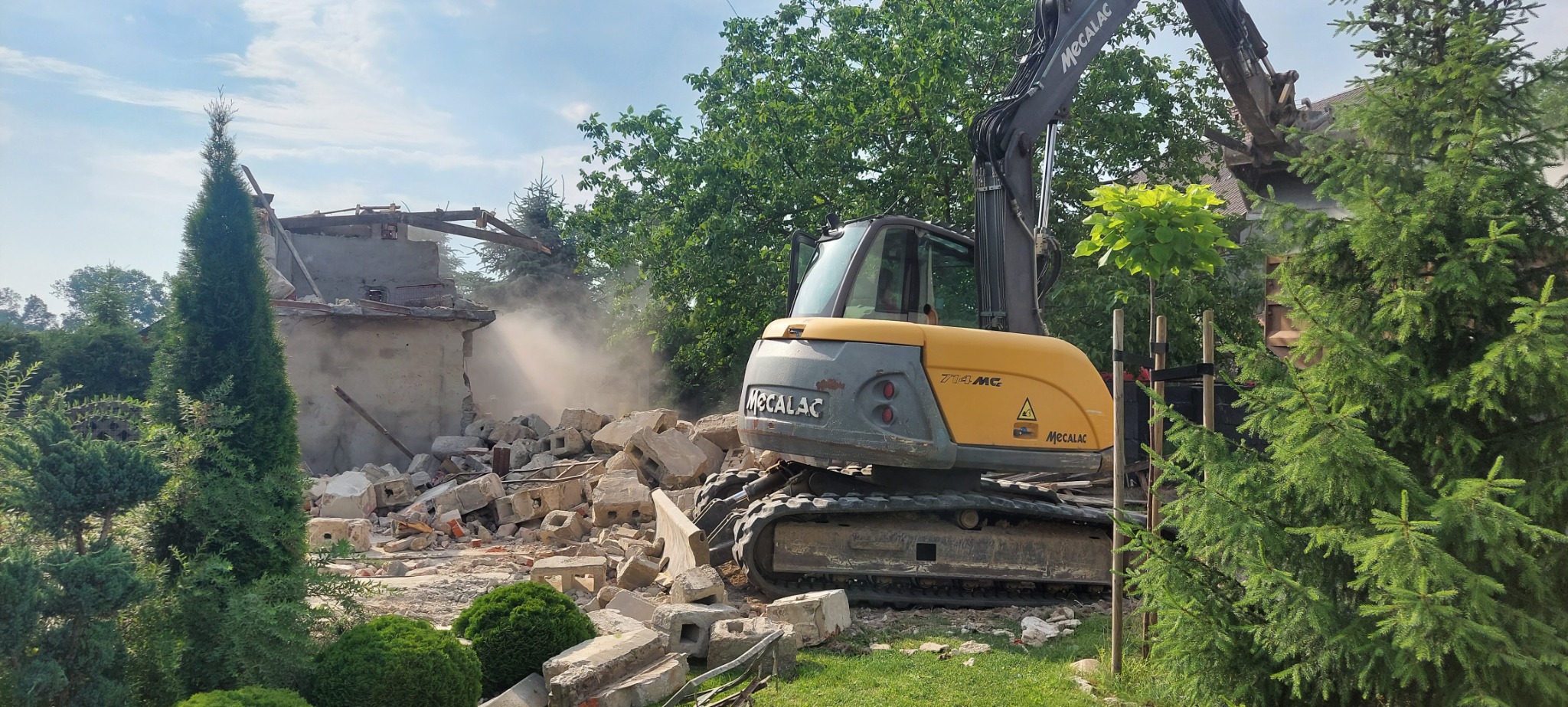 Żółta koparka Mecalac 714MC podczas wyburzania budynku z cegły, z widocznym pyłem i gruzem. W tle zieleń drzew i krzewów. Częściowo zniszczony budynek.