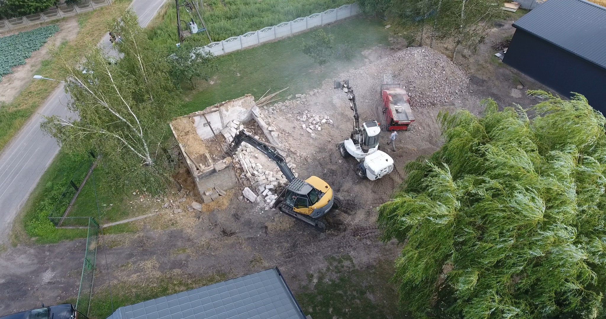 Z góry widok na plac budowy z żółtą koparką i białą maszyną do kruszenia gruzu podczas rozbiórki murowanego budynku. W tle hałda gruzu i zieleń.
