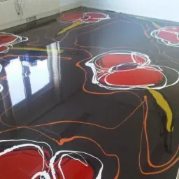 Żywiczna posadzka z abstrakcyjnym wzorem czerwonych kwiatów i pomarańczowych linii, odbijająca światło okna, tworząca unikatową, błyszczącą powierzchnię.