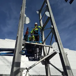 Dw&oacute;ch pracownik&oacute;w w podnośniku koszowym montuje elementy stalowej konstrukcji na tle błękitnego nieba i elewacji budynku.