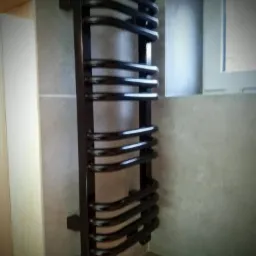 Czarny grzejnik łazienkowy drabinkowy, zamontowany na szarej ścianie z płytek, widoczny fragment okna i drzwi.