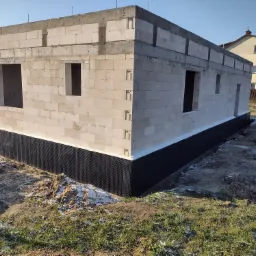 Surowy stan domu z betonu komórkowego z widoczną hydroizolacją fundamentów i otworami okiennymi, w tle widoczny inny budynek.