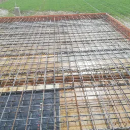 Zbrojenie płyty fundamentowej z siatki stalowej, drewniane szalunki i ceglana obudowa na obrzeżach, widok z góry w trakcie budowy.