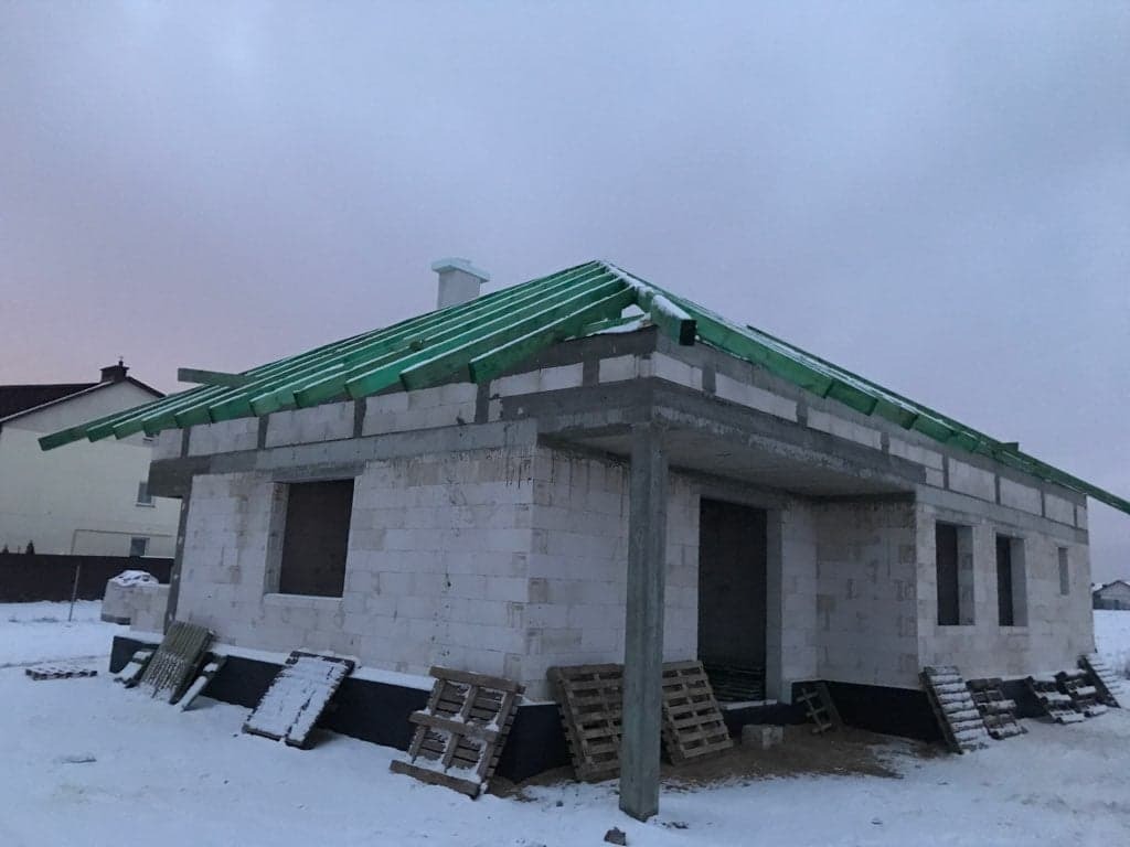 Surowy stan domu z białych bloczków w zimowej scenerii, widoczne zielone elementy konstrukcji dachu i palety pod śniegiem.