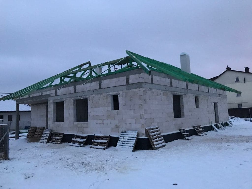 Niedokończony dom murowany w zimowej scenerii, widoczne palety pod oknami i zielona konstrukcja dachu w trakcie budowy.