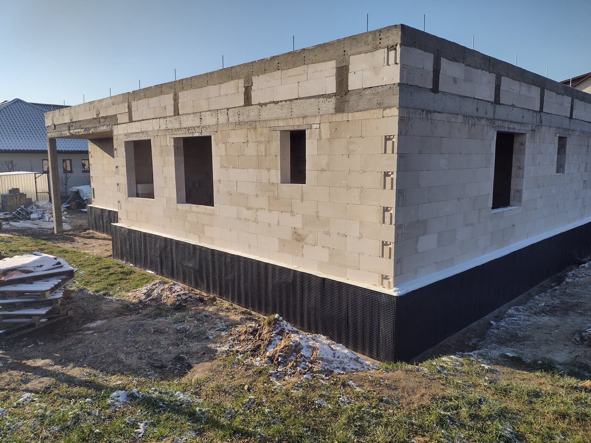 Surowy stan budynku z betonu komórkowego z widoczną izolacją fundamentów, otworami okiennymi i wystającymi prętami zbrojeniowymi w słoneczny dzień.