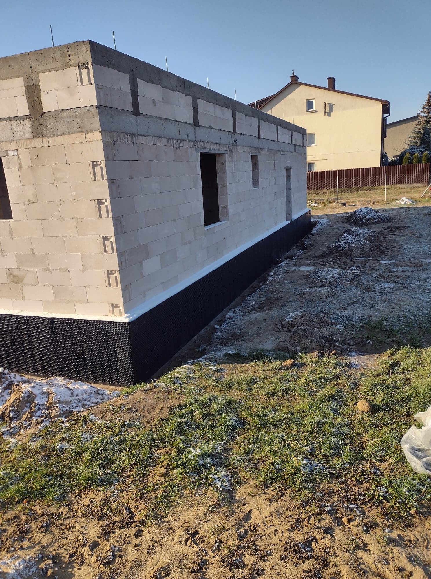 Wznoszenie ścian parterowego budynku z betonu komórkowego, widoczne okna i drzwi, izolacja fundamentów oraz zasypana ziemia wokół budynku.