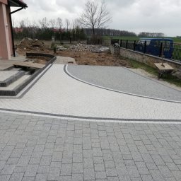 JACBRUK - Ułożona dwukolorowa nawierzchnia z kostki brukowej przed domem, widoczne schody wejściowe, przygotowane podłoże pod dalsze prace, w tle ogrodzenie panelowe i zabudowa.