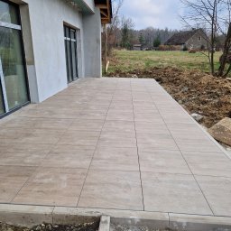 JACBRUK - Wyłożony płytkami taras przylegający do budynku, z widocznymi śladami stóp na powierzchni płytek i ziemią w tle.