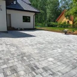 Ułożona szara kostka brukowa na podjeździe przed nowoczesnym domem z numerem 7B, w tle widoczna taczka i betoniarka.