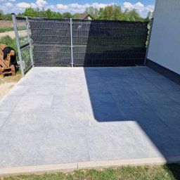 JACBRUK - Nowoczesny taras z szarych płyt kamiennych, obrzeża z jasnego betonu, ogrodzenie panelowe z czarną siatką, widoczny fragment białej ściany budynku i palety z drewnianymi meblami w tle.