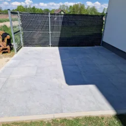 Nowoczesny taras z szarych płyt kamiennych, obrzeża z jasnego betonu, ogrodzenie panelowe z czarną siatką, widoczny fragment białej ściany budynku i palety z drewnianymi meblami w tle.