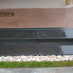 Nowoczesne, ciemne kamienne schody zewnętrzne z dwoma stopniami, wykończone białymi kamykami i trawą, prowadzące na taras z kostki brukowej w odcieniach brązu.