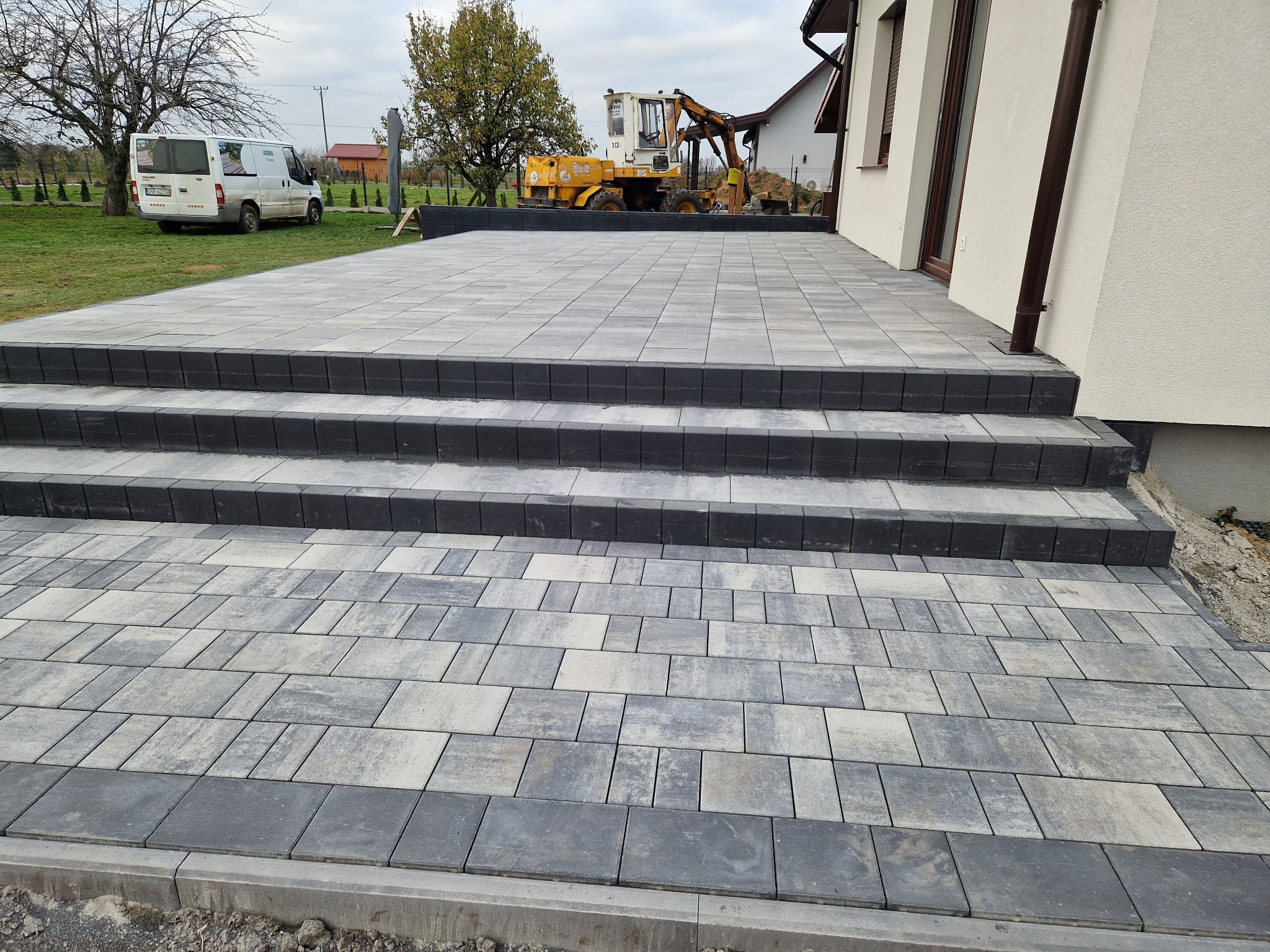 Nowy taras z szaro-beżowej kostki brukowej prowadzący do domu, z trzema stopniami wykończonymi ciemnoszarą kostką. W tle widoczny biały bus i żółta koparka.