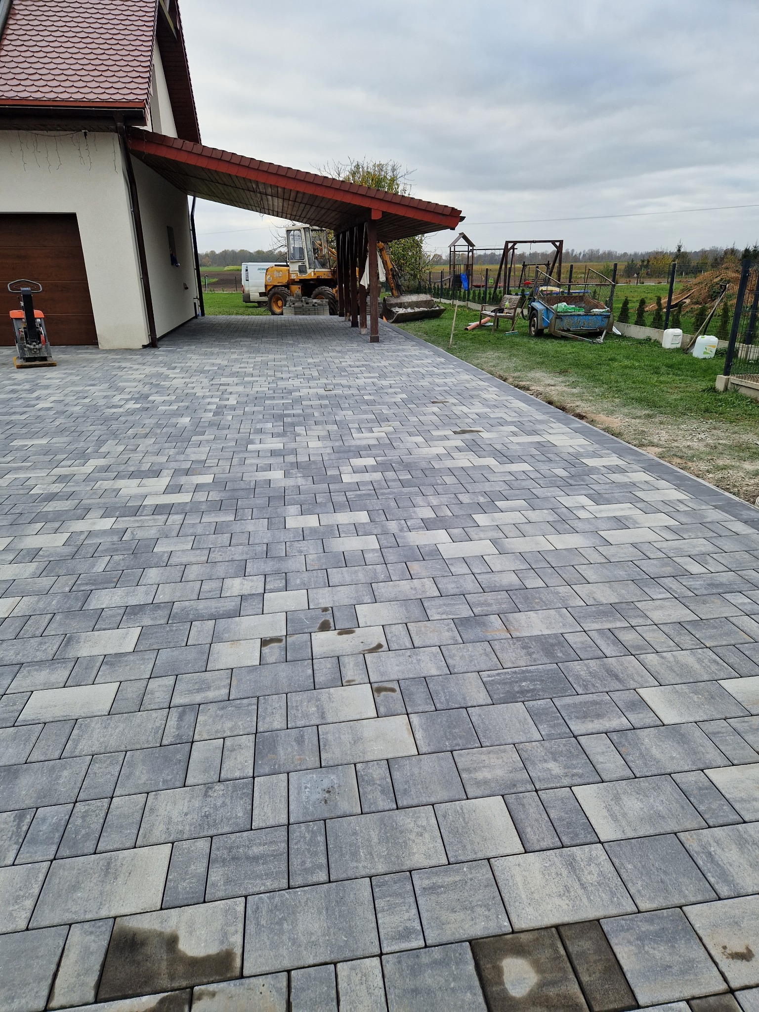 Ułożona szara kostka brukowa przed domem z podjazdem, widoczny sprzęt budowlany i przygotowany teren pod trawnik.