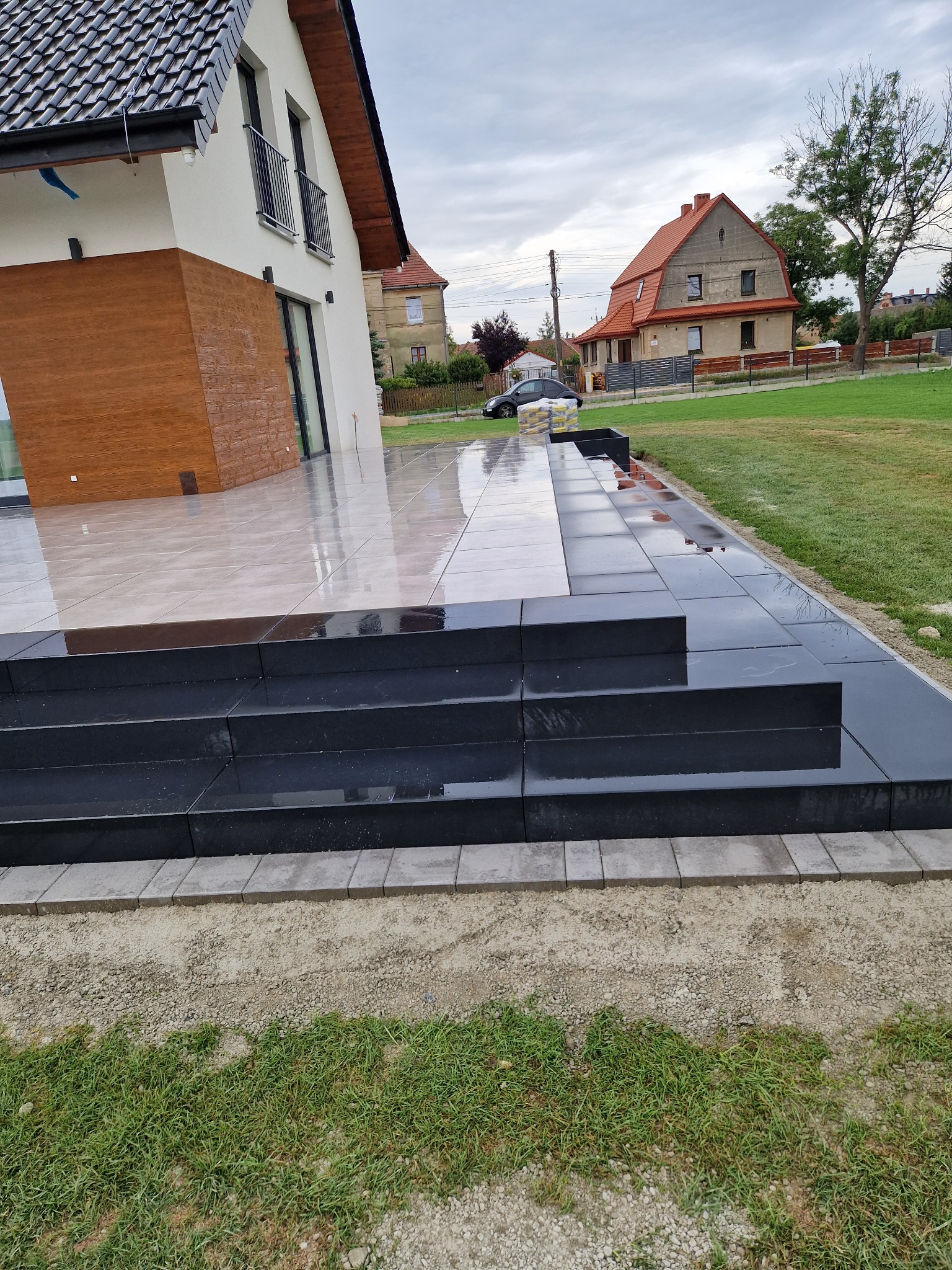 Nowoczesny taras z jasnych płytek przechodzący w czarne, kamienne schody, obrzeża z szarej kostki, widok na dom z drewnianą elewacją i trawnik