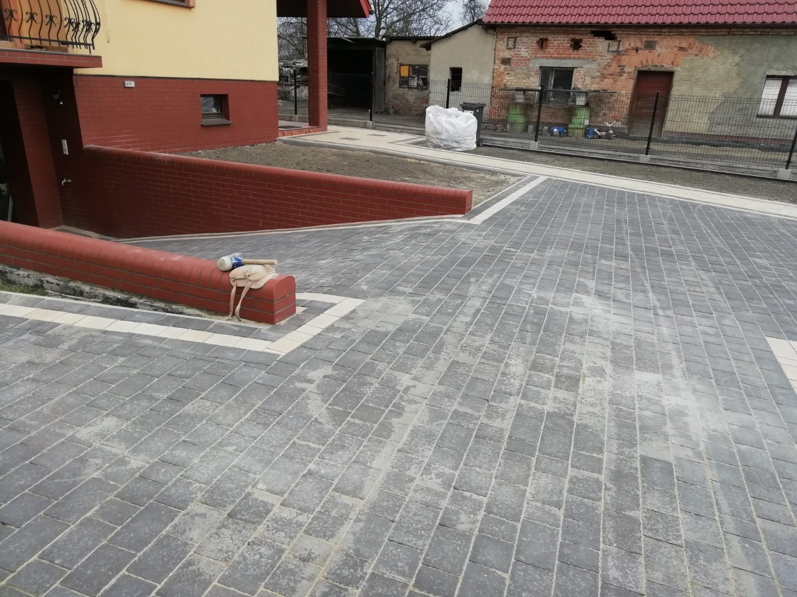 Ułożona szara kostka brukowa z jasnymi obramowaniami, widoczna torba narzędziowa i gumowy młotek na niskim murku z czerwonej cegły, dom z czerwono-żółtą elewacją w tle.