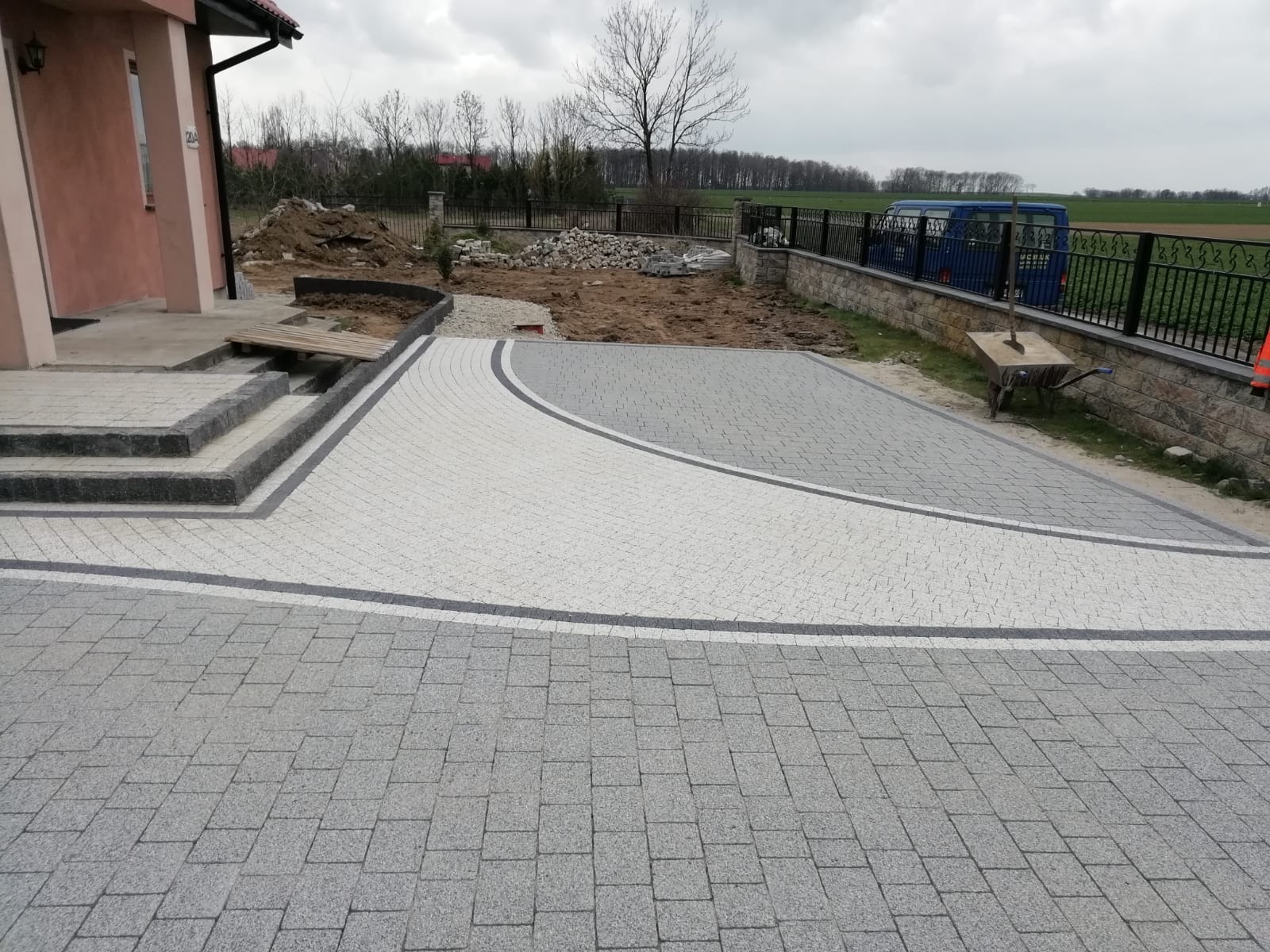 Ułożona dwukolorowa nawierzchnia z kostki brukowej przed domem, widoczne schody wejściowe, przygotowane podłoże pod dalsze prace, w tle ogrodzenie panelowe i zabudowa.