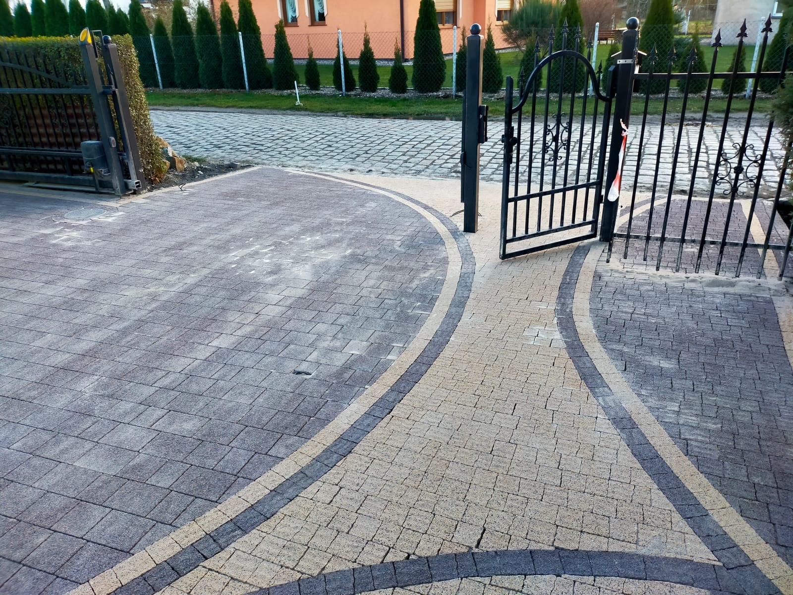 Ułożona kostka brukowa w dwóch odcieniach szarości i beżu tworząca wzór na podjeździe z otwartą, metalową bramą w tle.