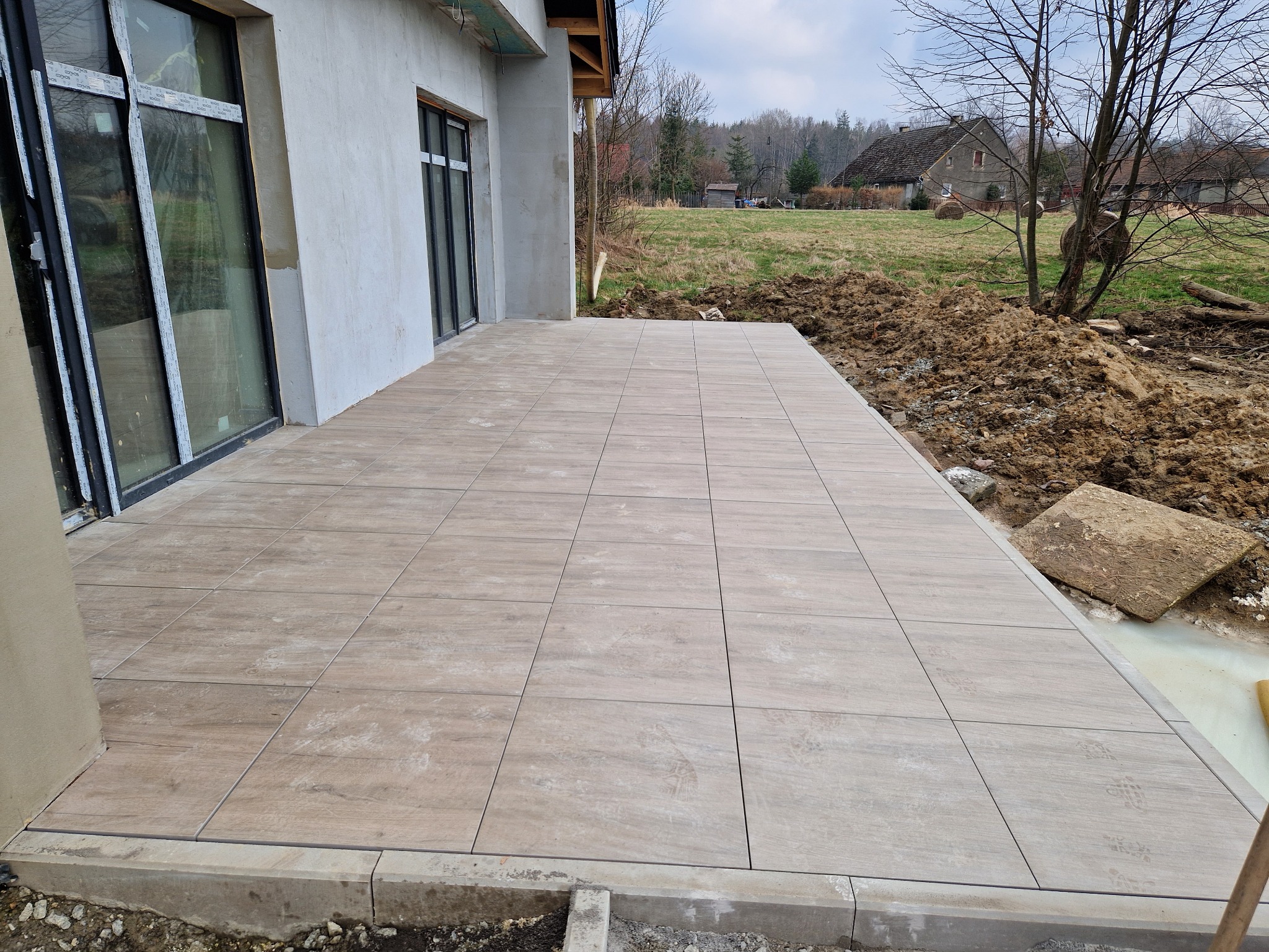 Wyłożony płytkami taras przylegający do budynku, z widocznymi śladami stóp na powierzchni płytek i ziemią w tle.