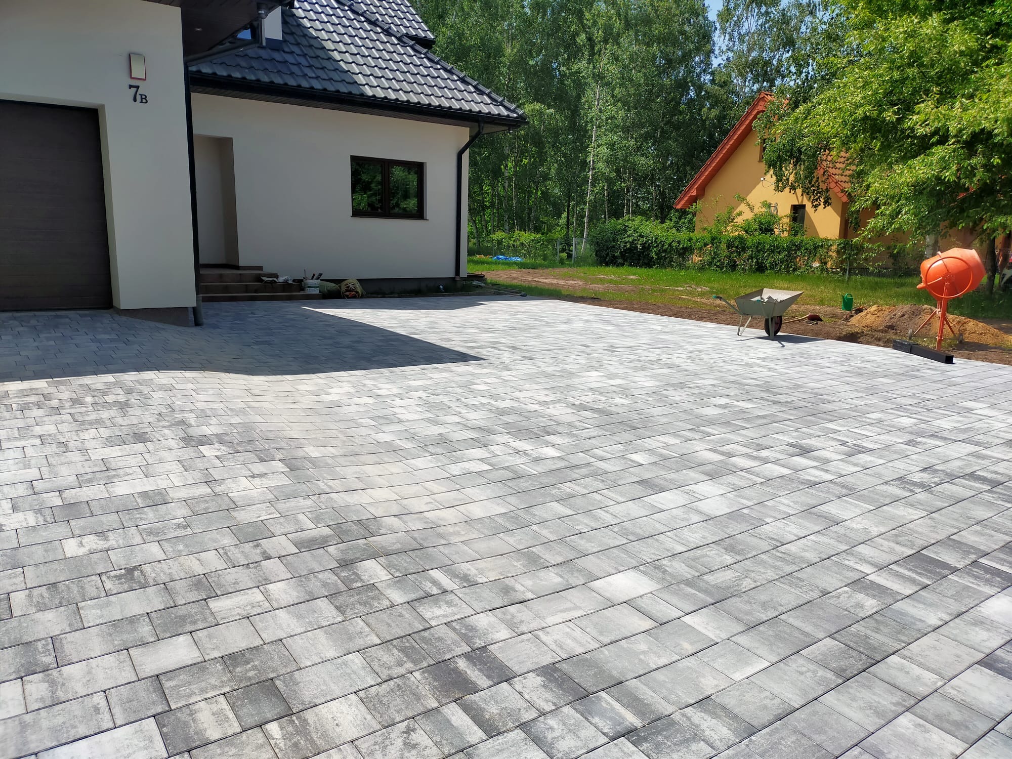 Ułożona szara kostka brukowa na podjeździe przed nowoczesnym domem z numerem 7B, w tle widoczna taczka i betoniarka.