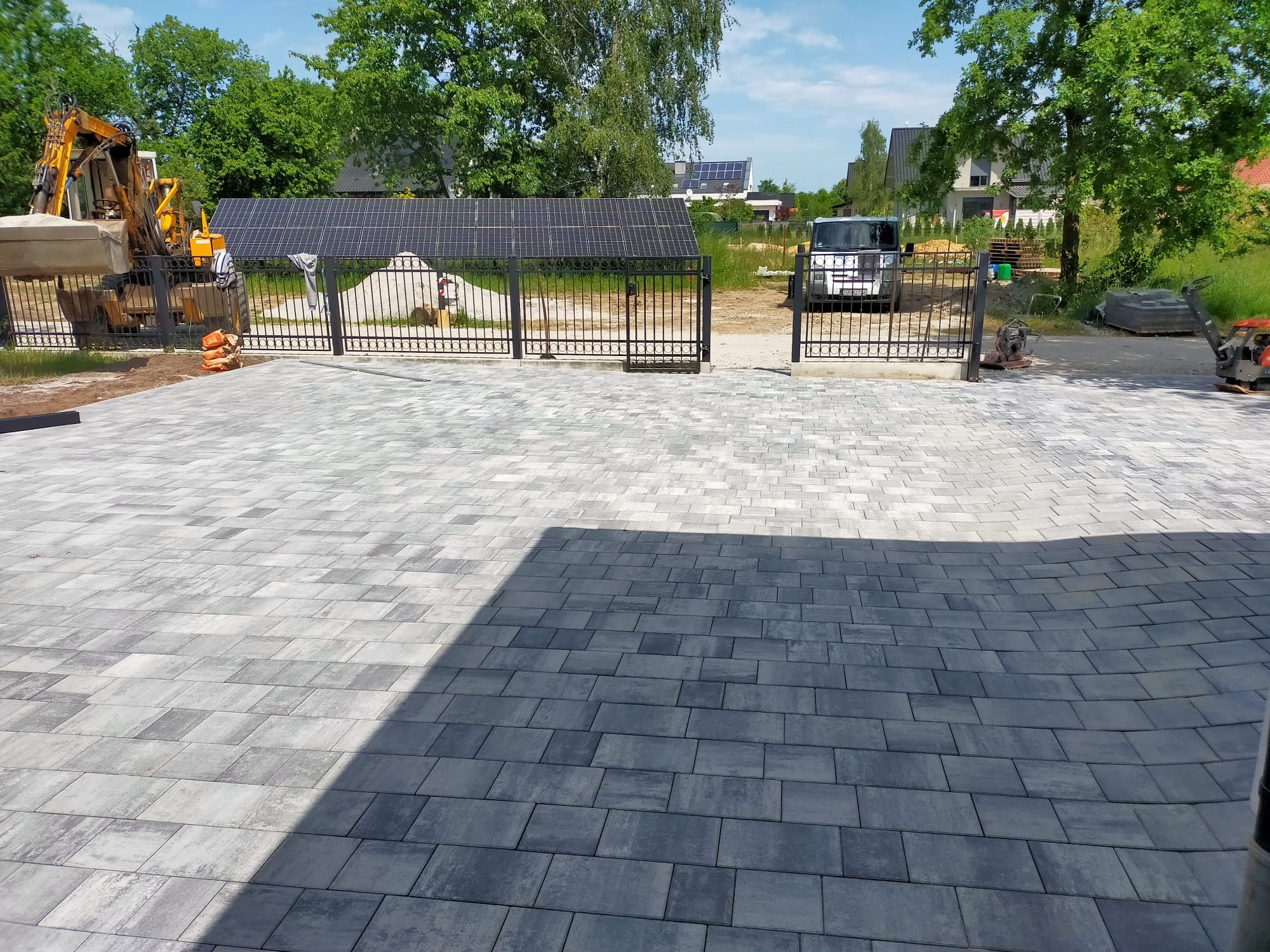 Ułożona szara kostka brukowa na podjeździe, widoczna brama panelowa z czarnymi przęsłami, koparka i ubijarka w tle, panele słoneczne na dachu budynku.