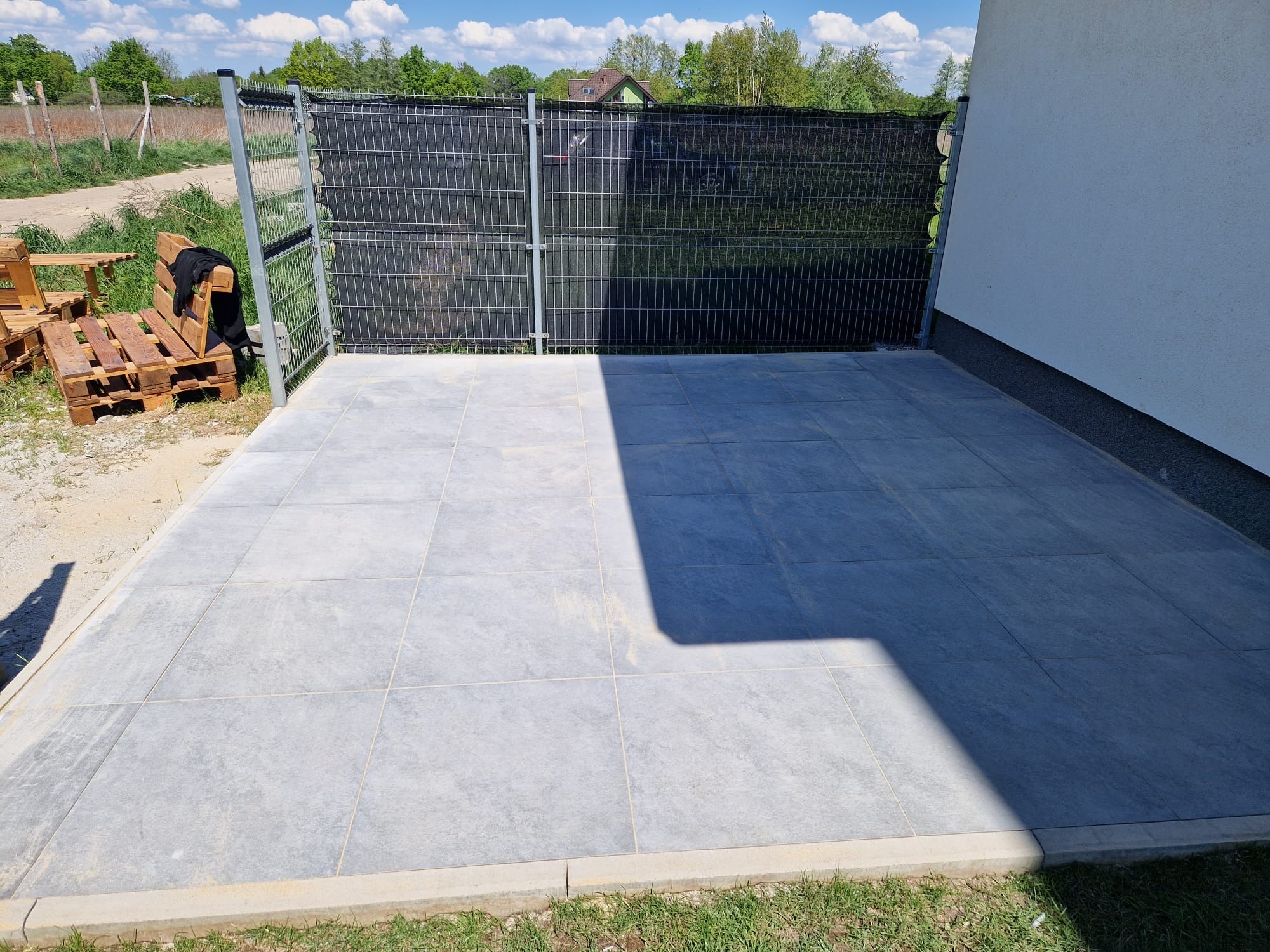 Nowoczesny taras z szarych płyt kamiennych, obrzeża z jasnego betonu, ogrodzenie panelowe z czarną siatką, widoczny fragment białej ściany budynku i palety z drewnianymi meblami w tle.