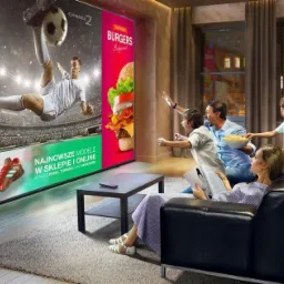 Rodzina ogląda duży ekran telewizora wyświetlającego mecz piłki nożnej i reklamę burgerów, z emocjami kibicując, podczas gdy mama czyta gazetę.