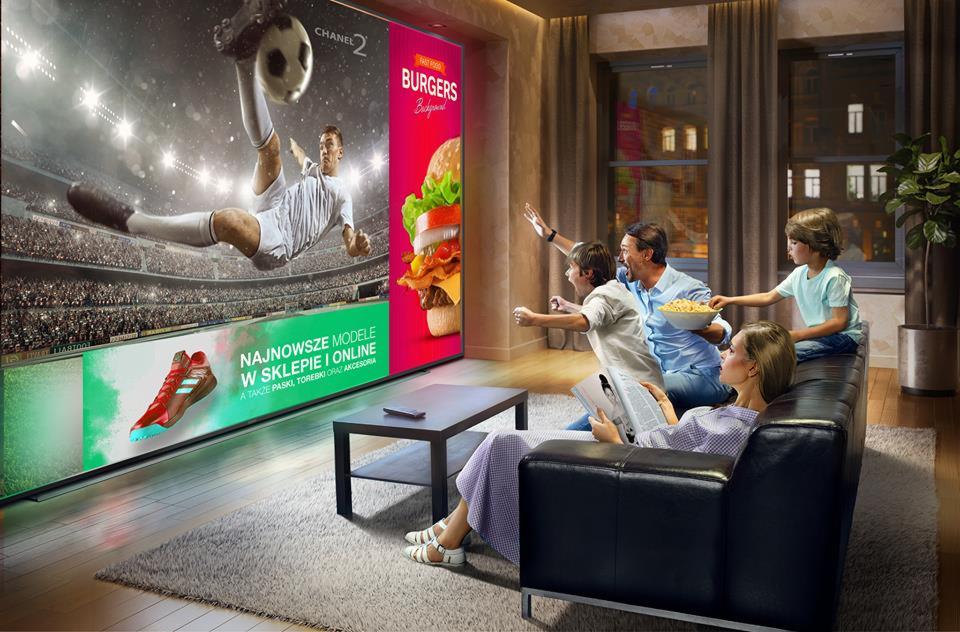 Rodzina ogląda duży ekran telewizora wyświetlającego mecz piłki nożnej i reklamę burgerów, z emocjami kibicując, podczas gdy mama czyta gazetę.